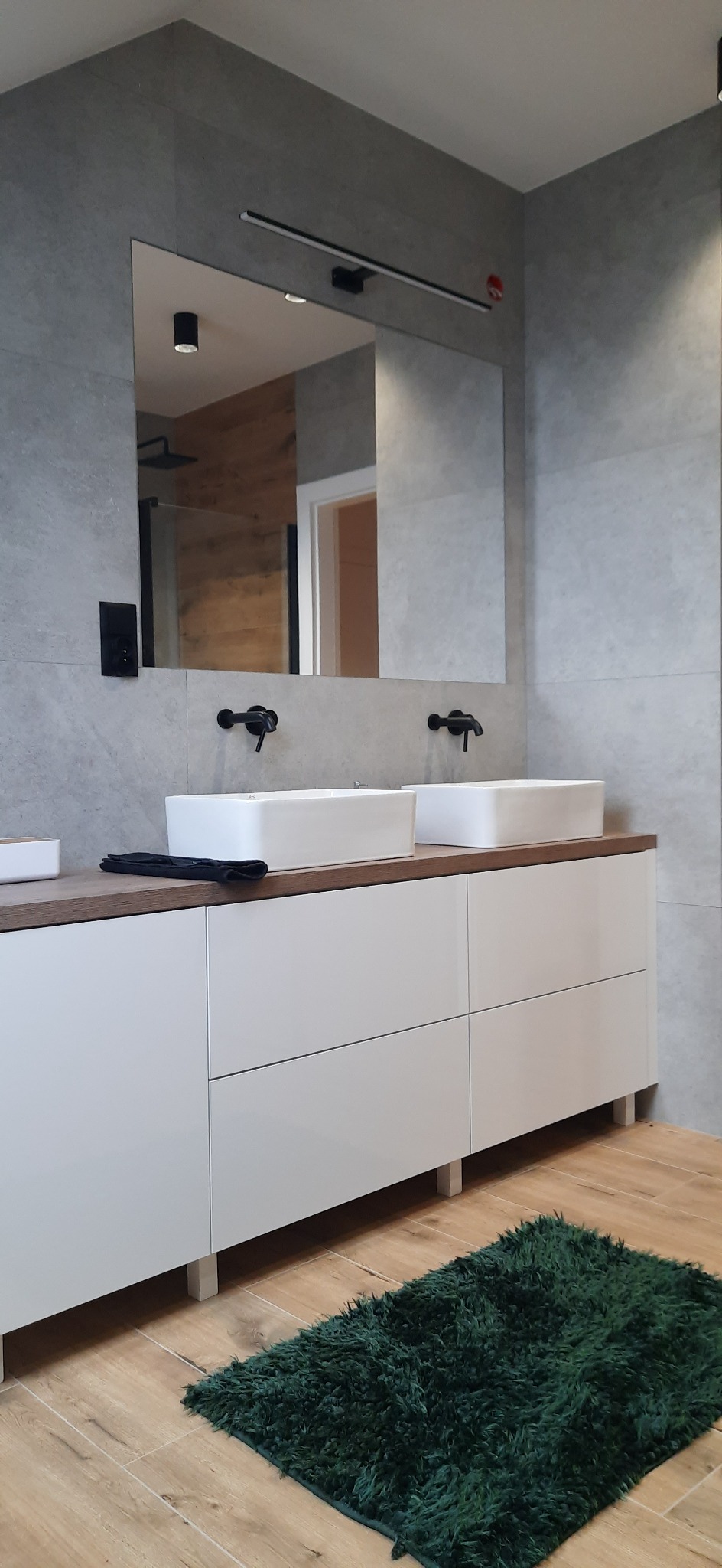 Nowoczesna łazienka z dwoma kwadratowymi umywalkami nablatowymi, czarną armaturą i szarą płytką imitującą beton, uzupełniona zielonym dywanikiem.