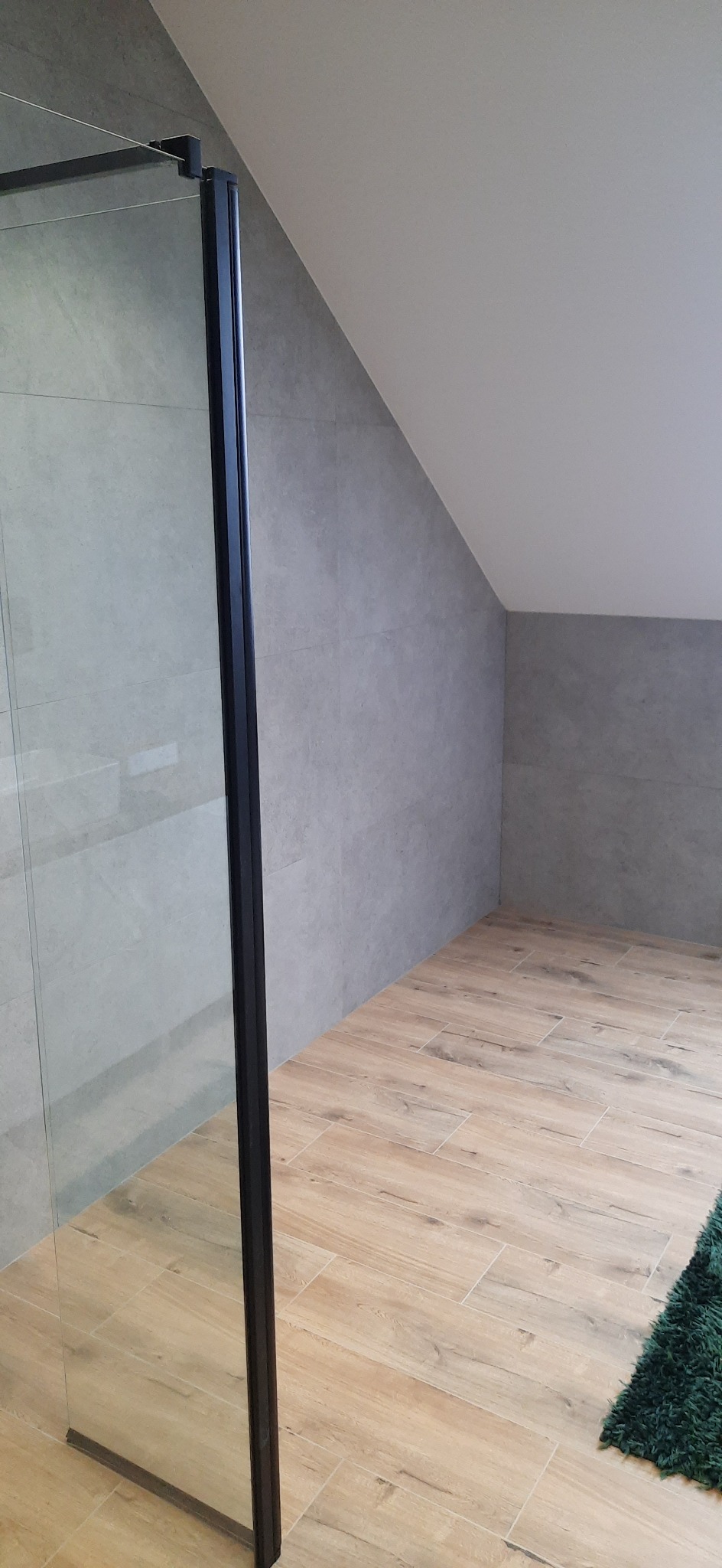 Wykończona łazienka na poddaszu z szarą płytką ścienną, drewnopodobną podłogą i kabiną prysznicową z czarną ramą oraz zielonym dywanikiem.