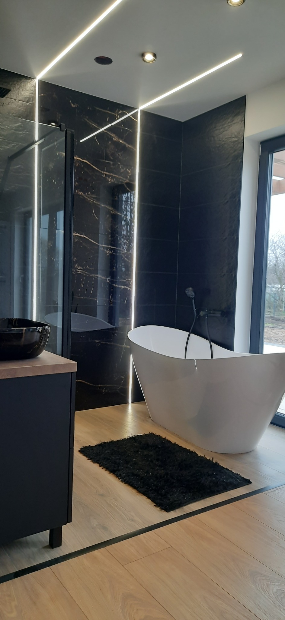 Nowoczesna łazienka z czarnymi płytkami imitującymi marmur, podświetleniem LED wzdłuż ścian, wolnostojącą białą wanną, czarną armaturą, drewnopodobną podłogą i czarnym dywanikiem.