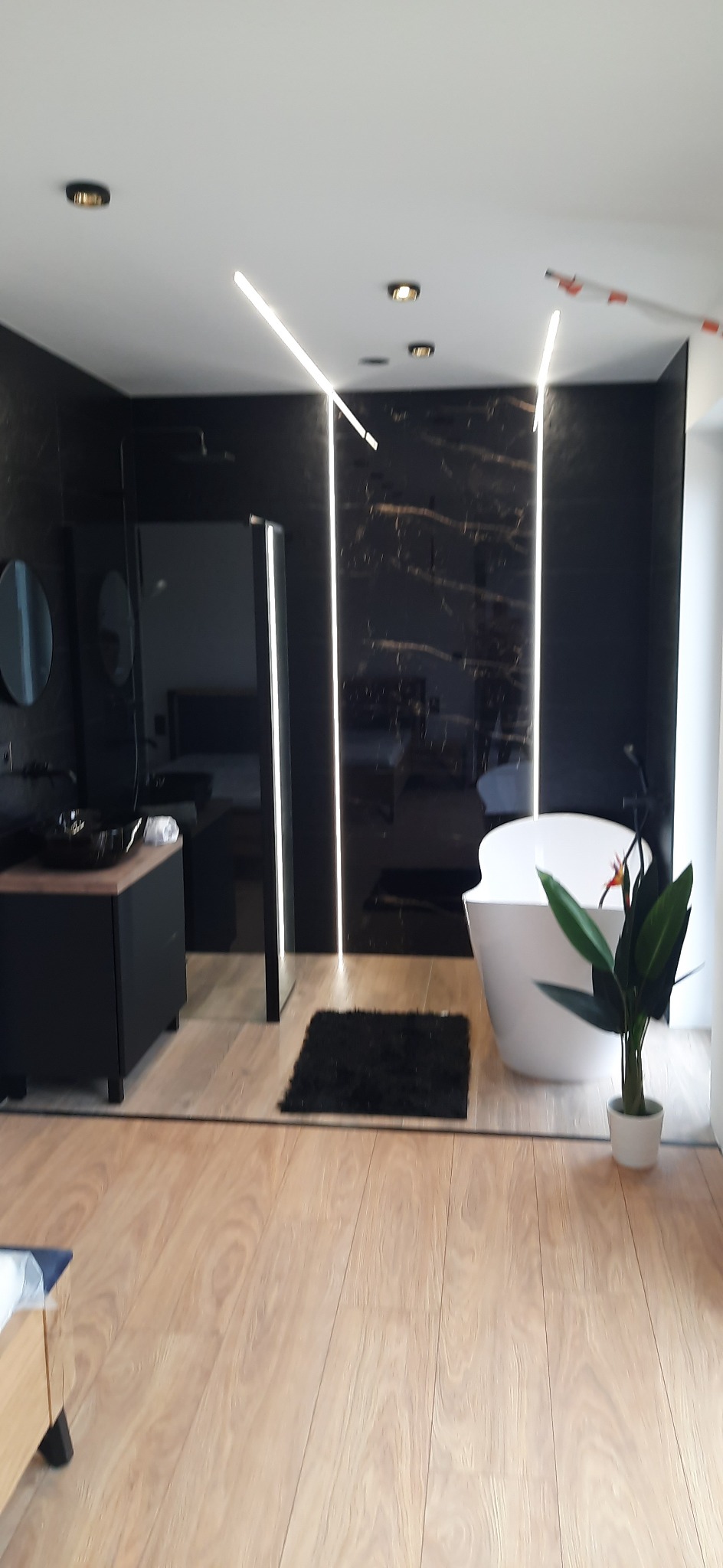 Nowoczesna łazienka z czarnymi marmurowymi płytkami, białym wolnostojącym zlewem i podświetlanymi liniami LED na ścianie, uzupełniona drewnopodobną podłogą i dekoracyjną rośliną w doniczce.