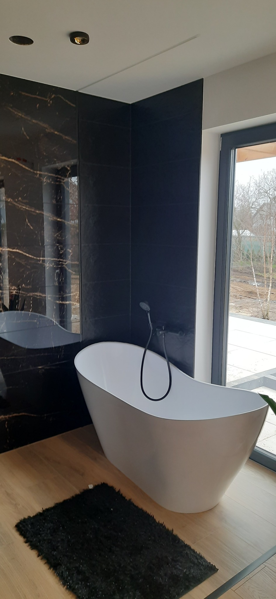 Nowoczesna łazienka z wolnostojącą białą wanną, czarną armaturą i płytkami imitującymi marmur z złotymi żyłami, drewnopodobna podłoga i czarny dywanik.
