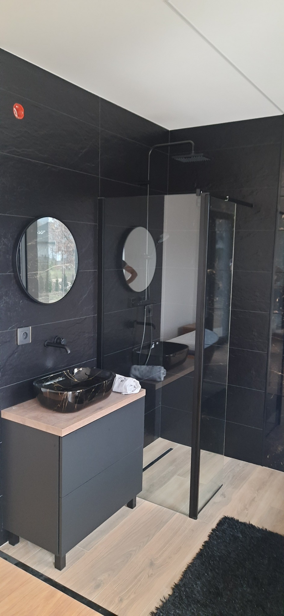 Nowoczesna łazienka z czarnymi płytkami, okrągłymi lustrami i kabiną prysznicową typu walk-in z czarnymi ramami, umywalka nablatowa z efektem marmuru na szafce z szufladami.