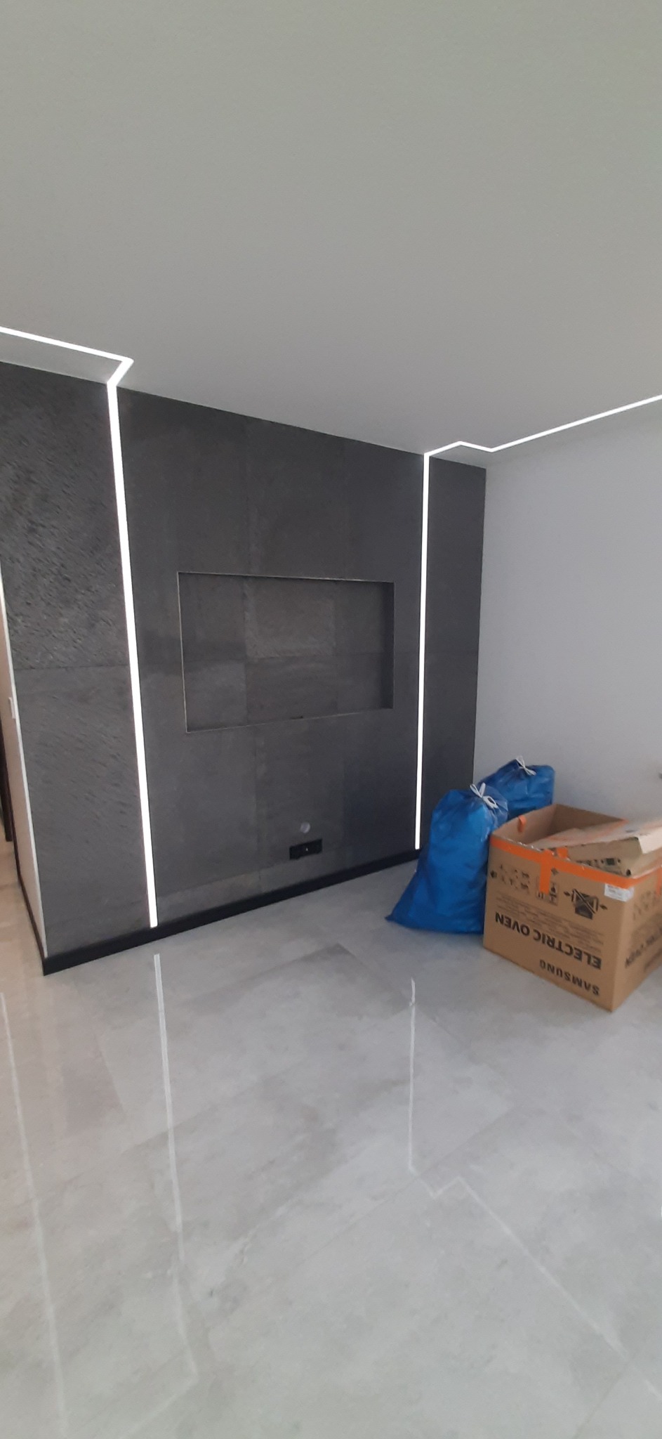 Szara ściana z wnęką i pionowym oświetleniem LED, marmurowa podłoga, karton po piekarniku i worki z odpadami w nowym mieszkaniu.