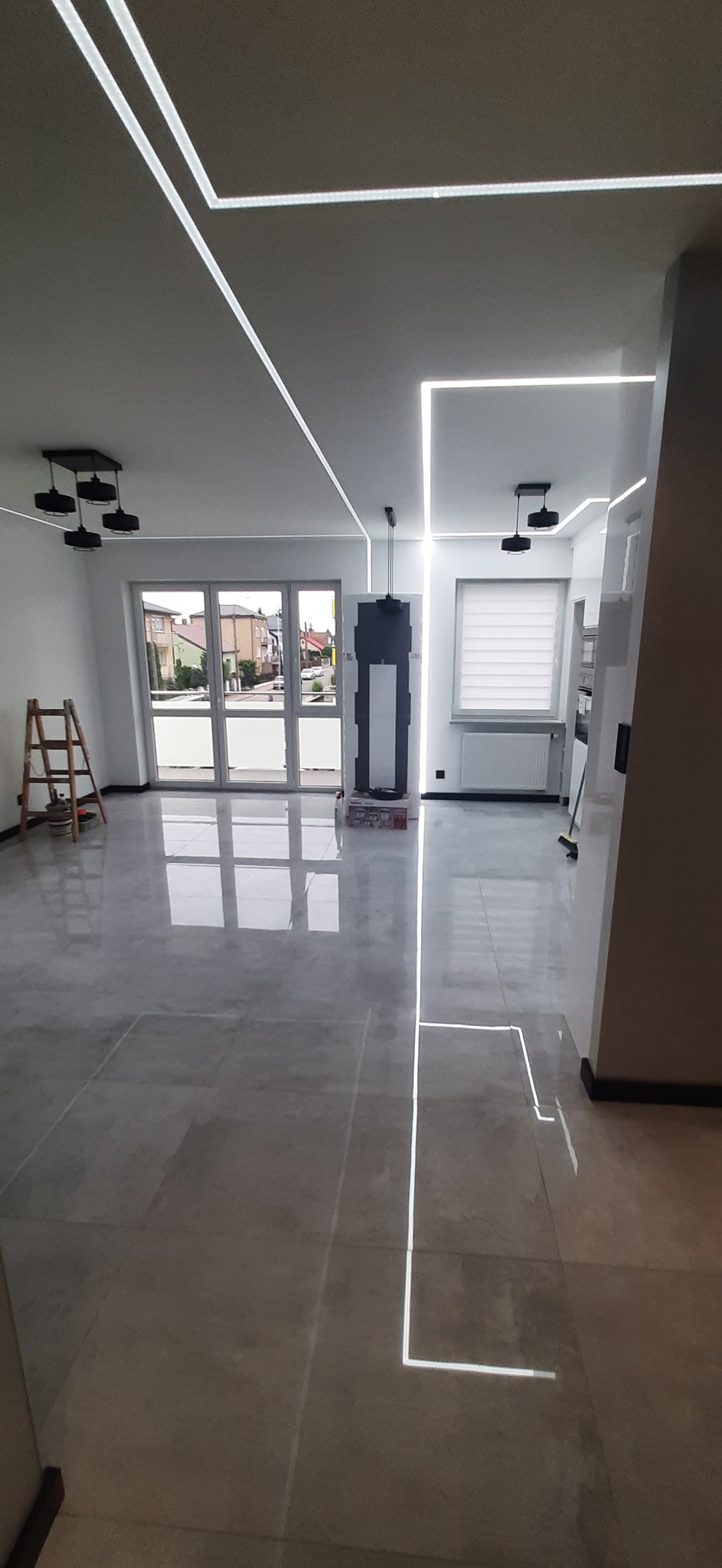 Salon po remoncie z podświetleniem LED w suficie, szare płytki podłogowe z połyskiem, drabina malarska i kartony w rogu pokoju.