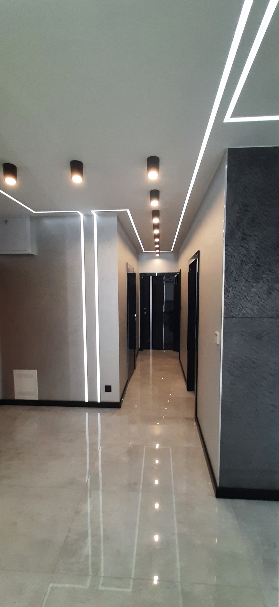 Nowoczesne oświetlenie LED w przedpokoju z szarą ścianą i marmurową posadzką, widoczne czarne drzwi w głębi korytarza.