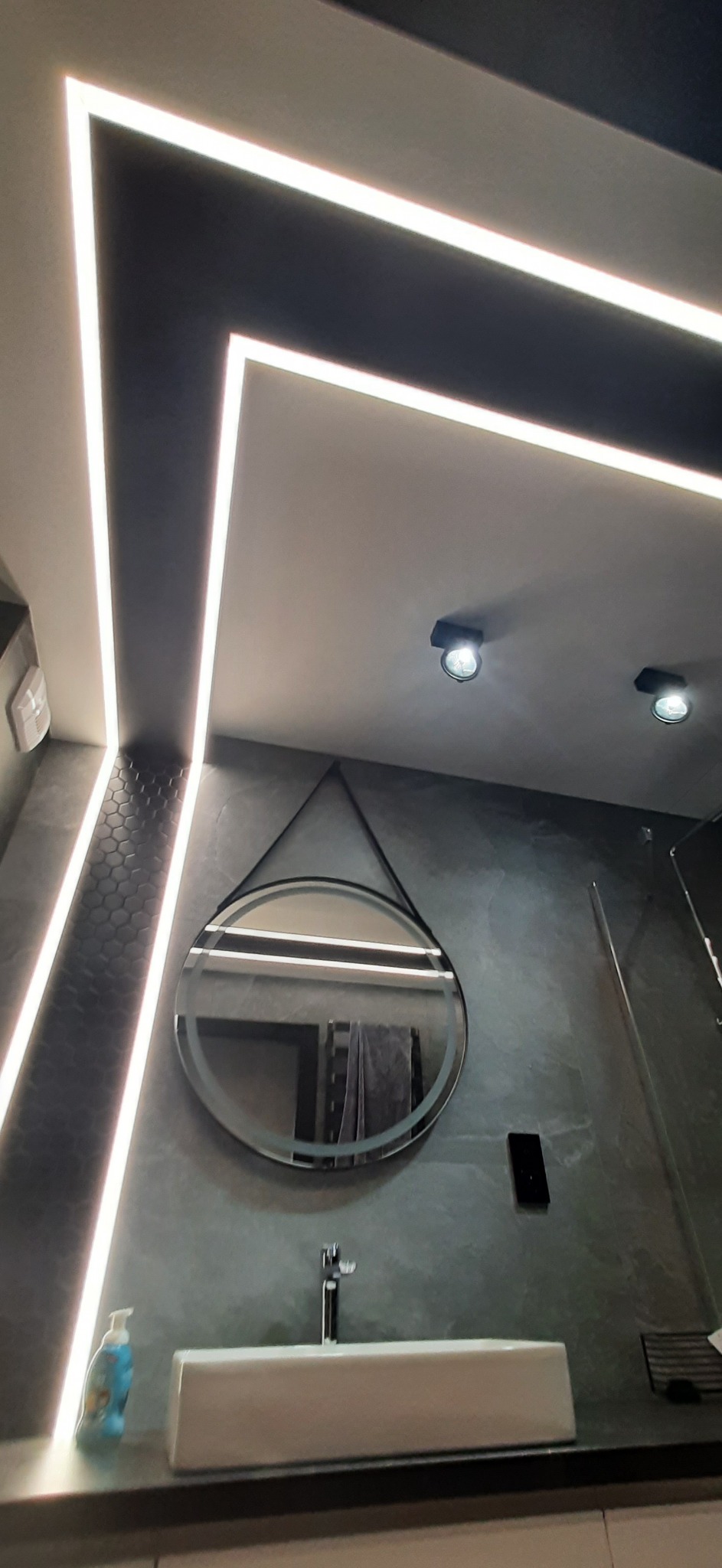 Nowoczesna łazienka z szarą, teksturowaną ścianą, okrągłym lustrem na pasku, białą umywalką i podświetleniem LED w suficie.