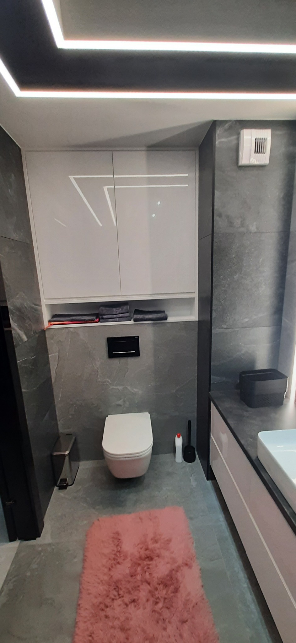 Nowoczesna łazienka z szarymi płytkami imitującymi kamień, podwieszaną toaletą, białymi szafkami z połyskiem, oświetleniem LED na suficie i różowym dywanikiem.