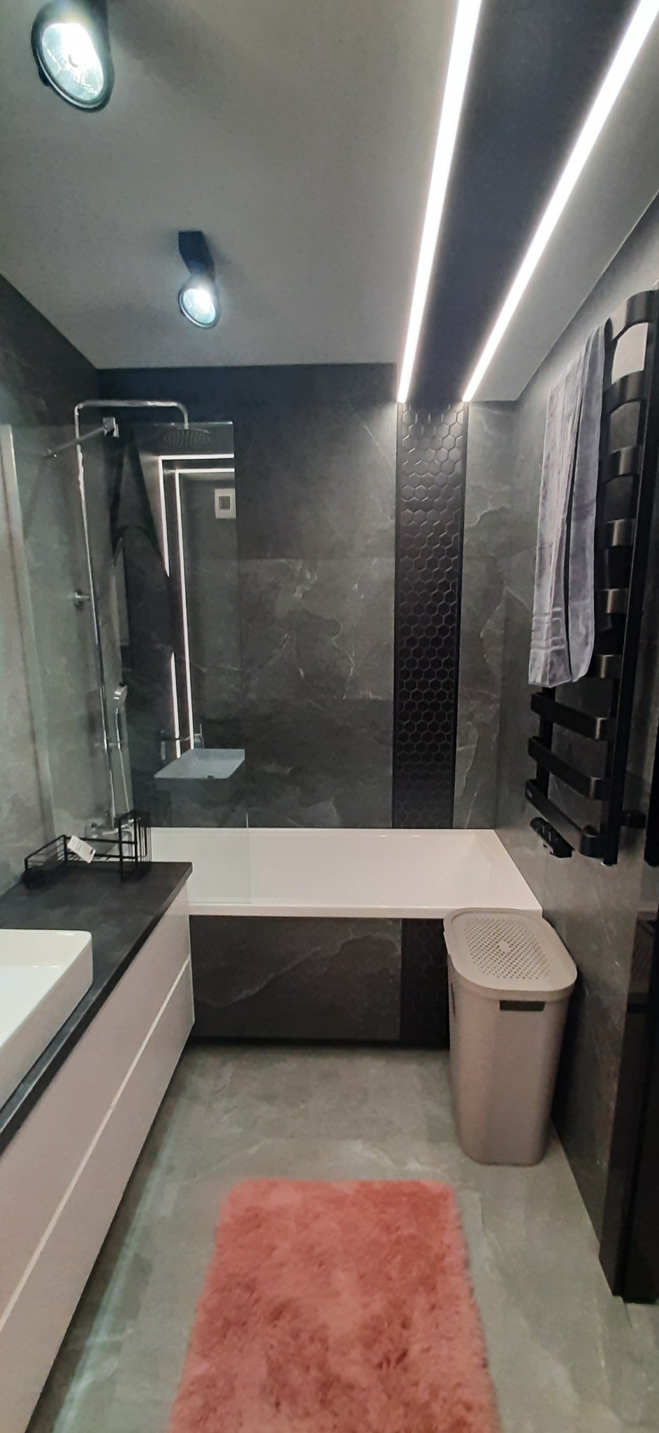 Nowoczesna łazienka z ciemnymi marmurowymi płytkami, podświetleniem LED, wanną, kabiną prysznicową i różowym dywanikiem.