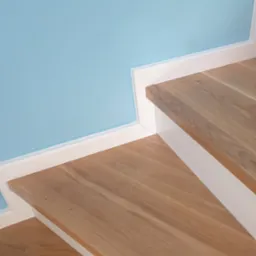 Fragment drewnianych schodów z białymi listwami przypodłogowymi na tle błękitnej ściany, widok z góry.