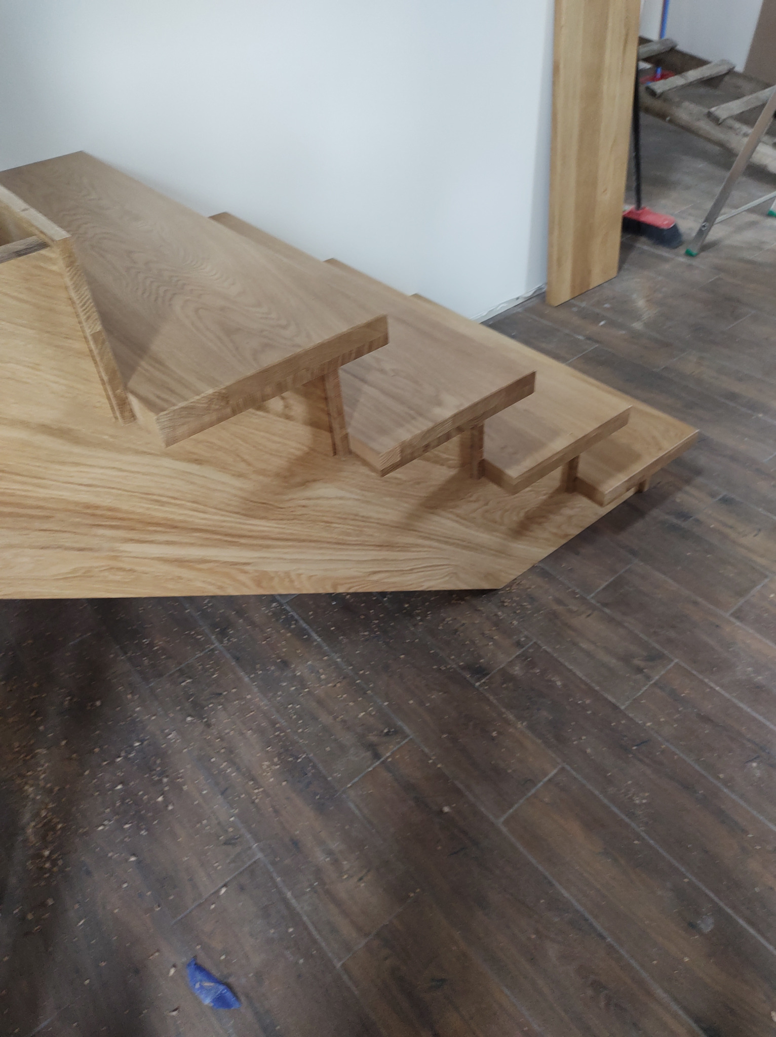 Surowe, drewniane stopnie schodów w trakcie montażu, widoczne drobne wióry na podłodze z płytek imitujących drewno, w tle ściana i fragment drabiny.