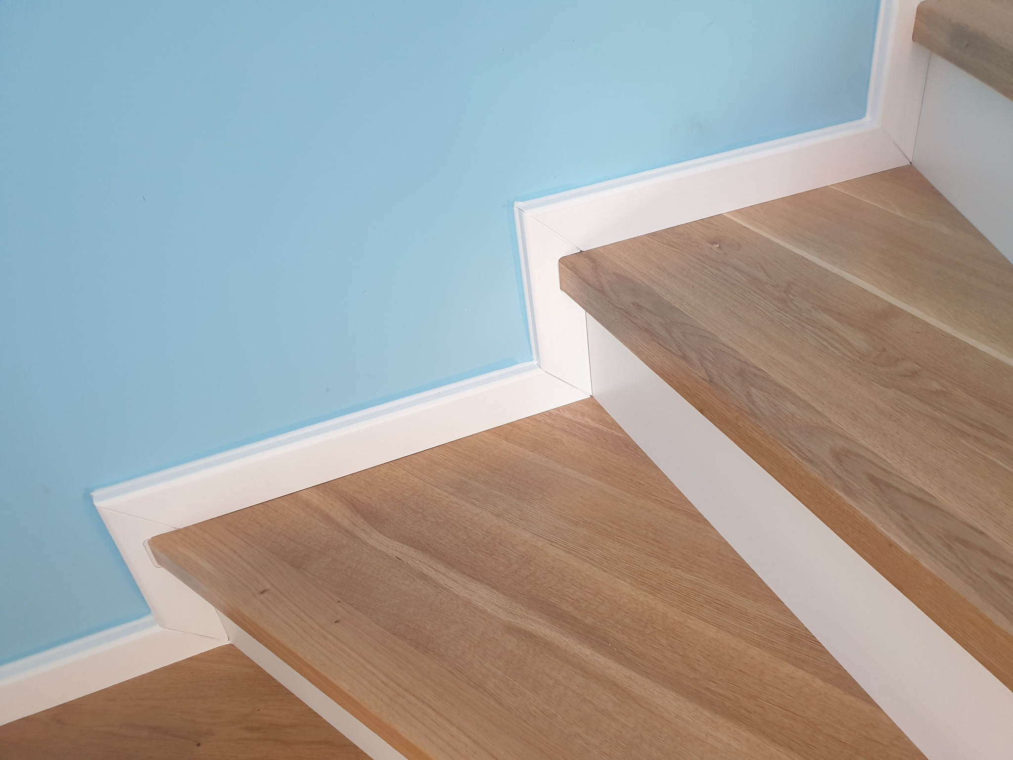 Fragment drewnianych schodów z białymi listwami przypodłogowymi na tle błękitnej ściany, widok z góry.