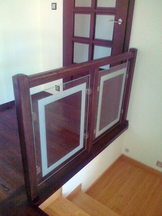 Szczegółowe ujęcie drewnianej balustrady schodowej z panelami szklanymi, widoczne metalowe mocowania szyb, drewniane schody i drzwi w tle.