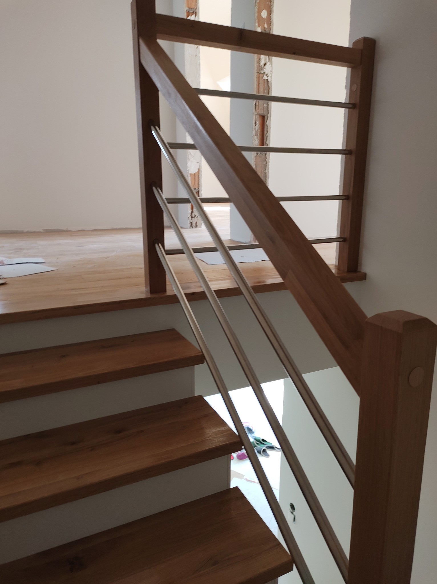 Nowoczesne schody z drewnianymi stopniami i balustradą z drewnianymi słupkami oraz metalowymi, poziomymi prętami, widoczne podczas montażu w jasnym wnętrzu.