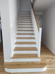 Jasne schody z drewnianymi stopniami i białymi podstopnicami oraz balustradą, prowadzące na górę w nowoczesnym wnętrzu. Widoczny fragment drewnianej podłogi i ściany w tle.