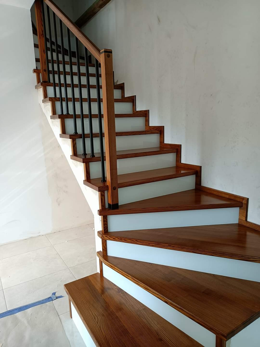 Nowoczesne schody z drewna i białych podstopnic, czarna metalowa balustrada, widoczne ślady prac wykończeniowych na ścianie i podłodze.