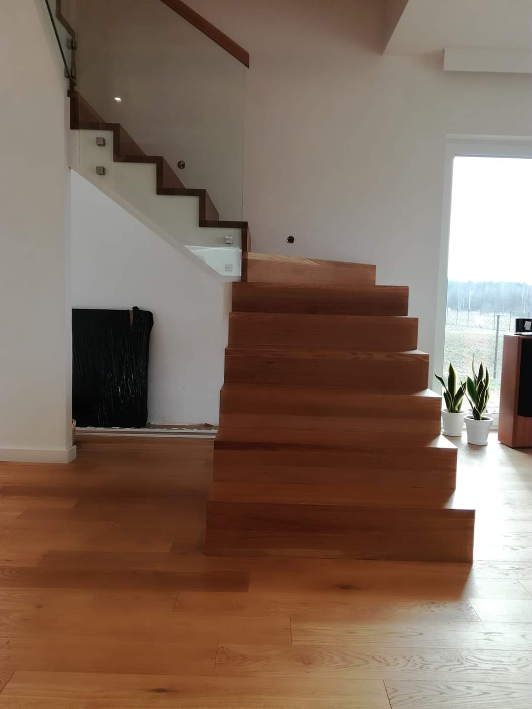 Nowoczesne schody z drewna z przezroczystą balustradą szklaną, oświetleniem punktowym i podłogą z desek w jasnym wnętrzu domu.