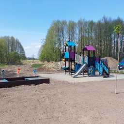 ARCHEMI Sp. z o.o. - Nowo wybudowany plac zabaw z kolorowymi konstrukcjami, piaskownicą z elementami wodnymi i huśtawkami sprężynowymi na tle lasu i błękitnego nieba, z widocznym samochodem dostawczym i pracownikiem...