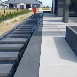 ARCHEMI Sp. z o.o. - Schody z kostki brukowej w trakcie budowy, widoczne warstwy pod kostką, obok ułożona jasna kostka brukowa, w tle budynek i osoba w czerwonym ubraniu.