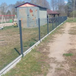 Panelowe ogrodzenie z podmurówką betonową, widok z boku na tle placu zabaw i zabudowań gospodarczych.