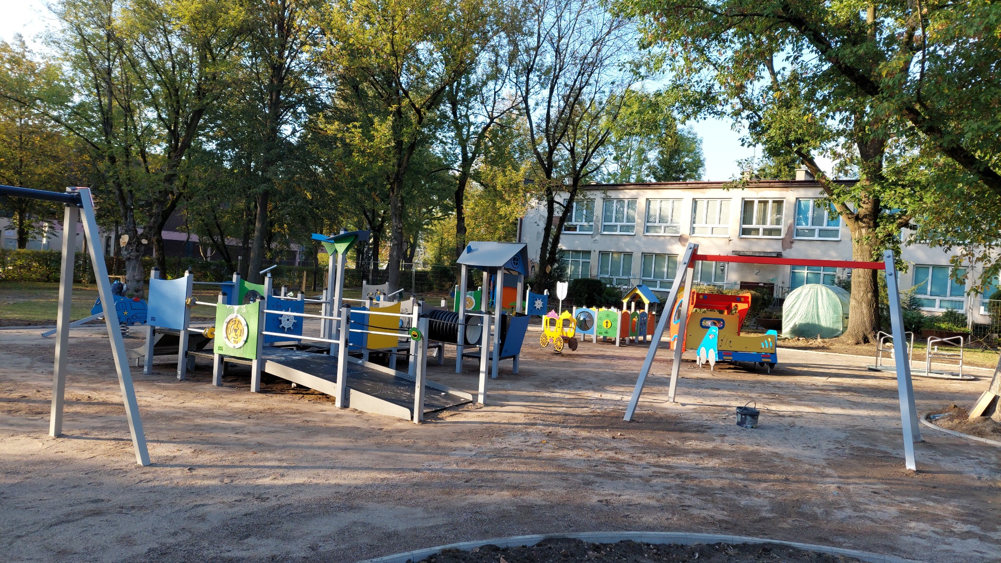 Kolorowy plac zabaw z huśtawkami i zjeżdżalnią na piaszczystym podłożu, na tle budynku szkoły i drzew.