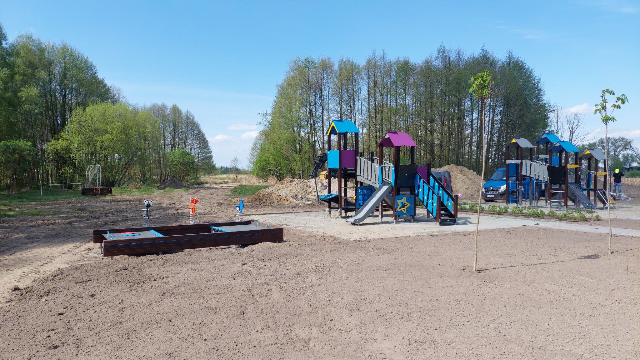 Nowo wybudowany plac zabaw z kolorowymi konstrukcjami, piaskownicą z elementami wodnymi i huśtawkami sprężynowymi na tle lasu i błękitnego nieba, z widocznym samochodem dostawczym i pracownikiem...