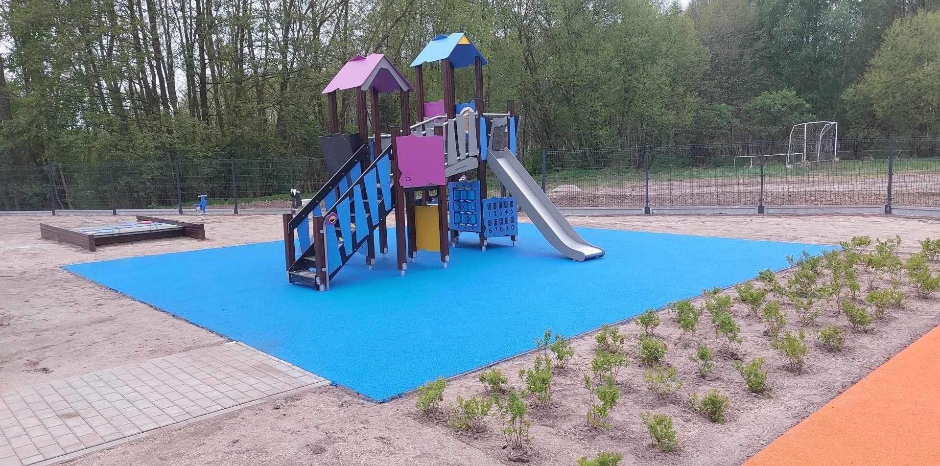 Kolorowy plac zabaw z wieżą, zjeżdżalnią, schodami i panelami edukacyjnymi, otoczony niebieską nawierzchnią amortyzującą i młodymi krzewami.