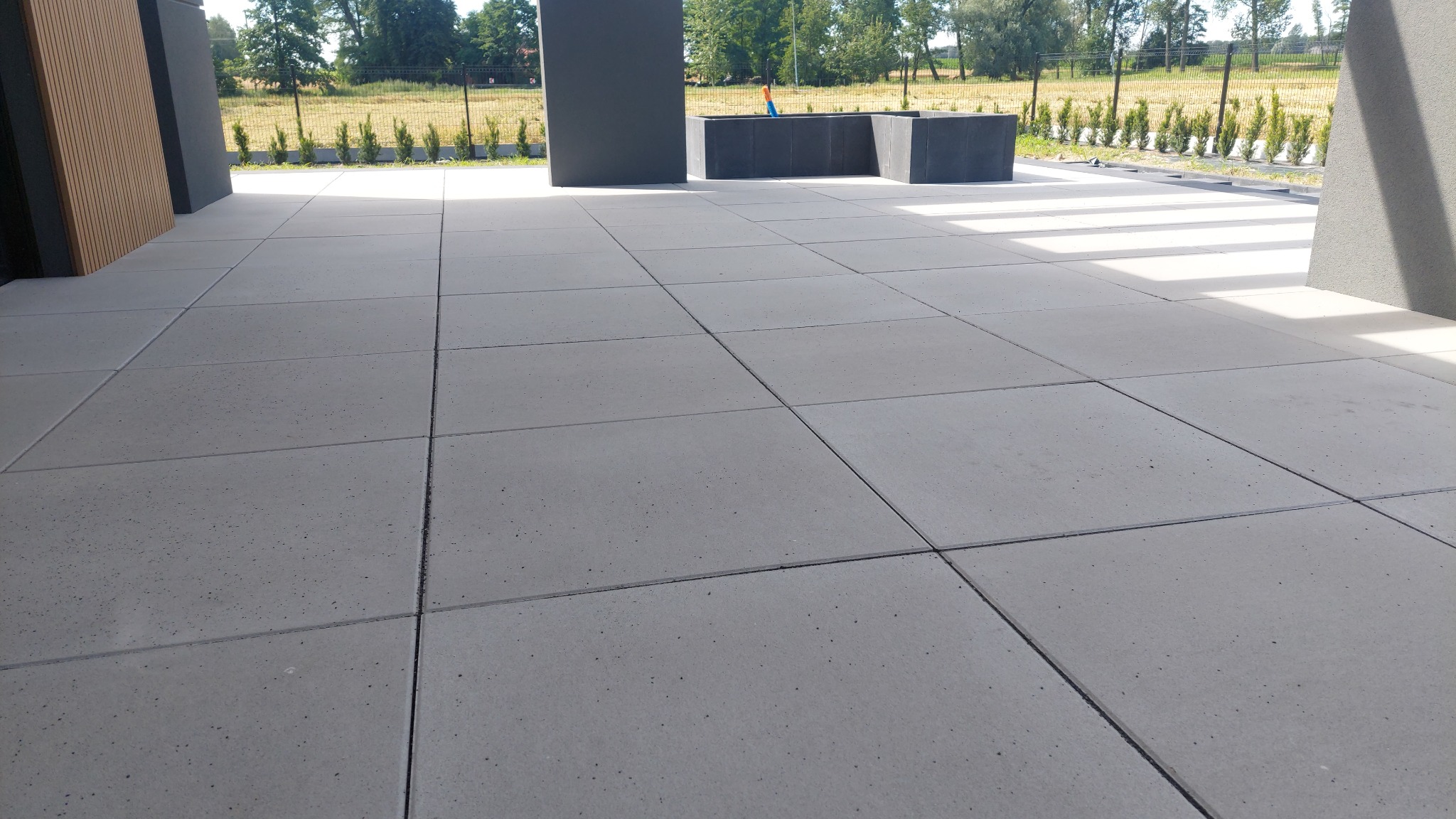 Nowoczesny taras wyłożony szarą kostką brukową o regularnym kształcie, z widocznymi donicami i roślinami w tle.