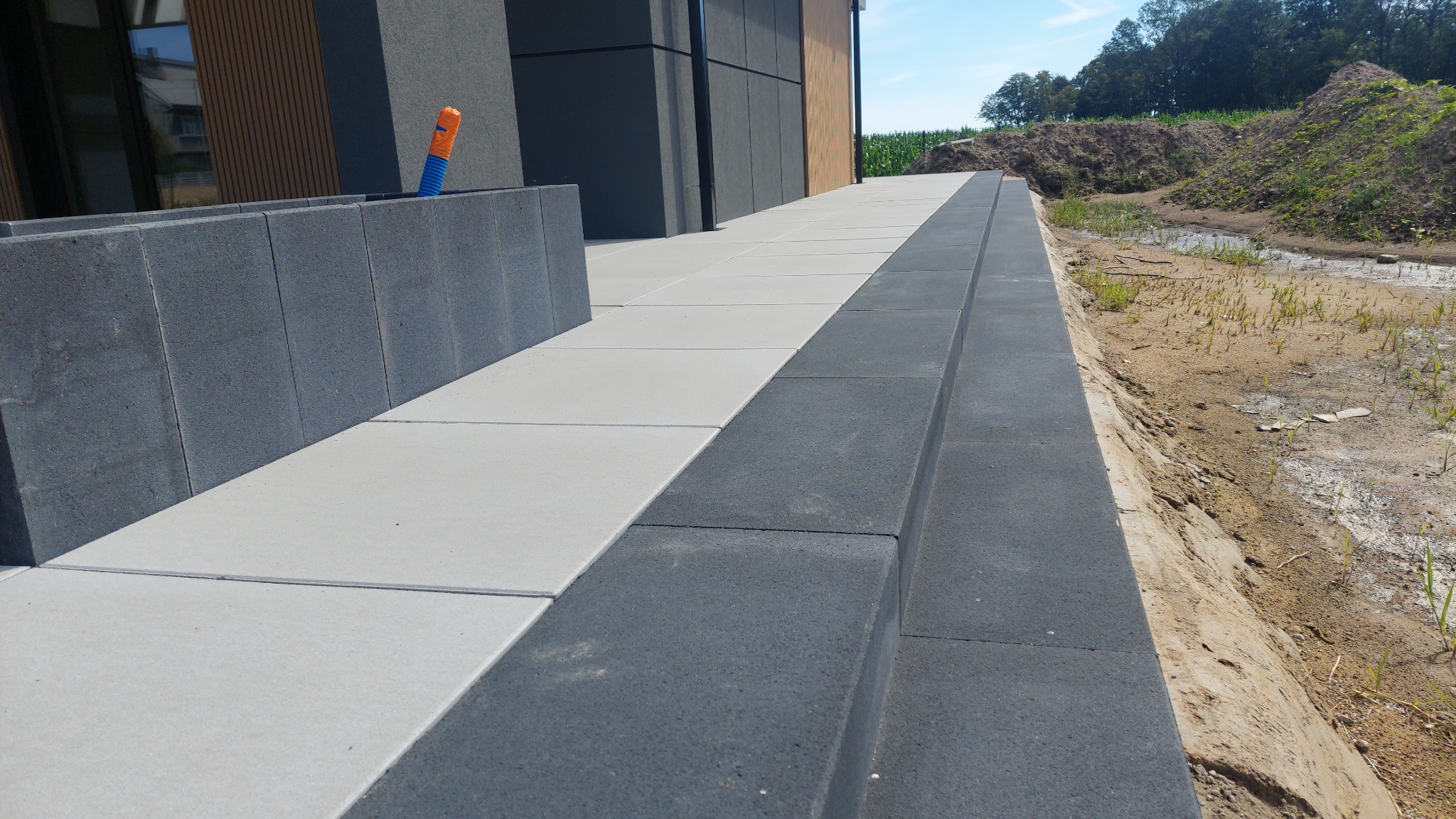 Jasnoszara kostka brukowa z ciemnoszarym obrzeżem, widoczna rura w ścianie, trawa w tle.