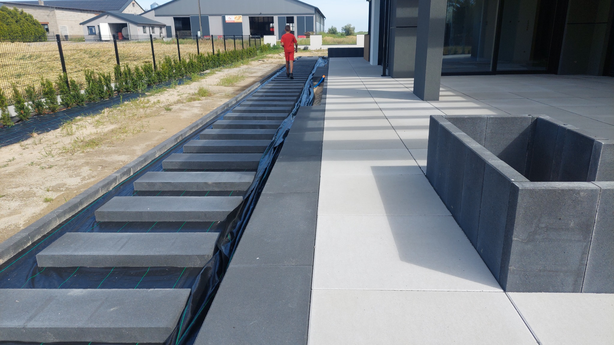 Schody z kostki brukowej w trakcie budowy, widoczne warstwy pod kostką, obok ułożona jasna kostka brukowa, w tle budynek i osoba w czerwonym ubraniu.