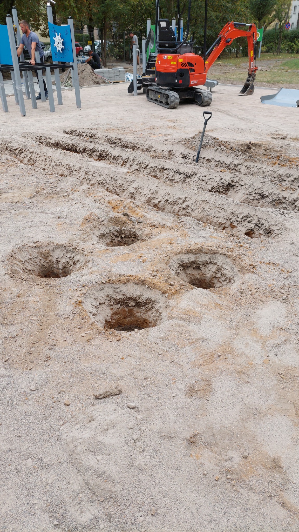 Przygotowanie terenu pod plac zabaw: wykopane doły, mini koparka i pracownik w tle.