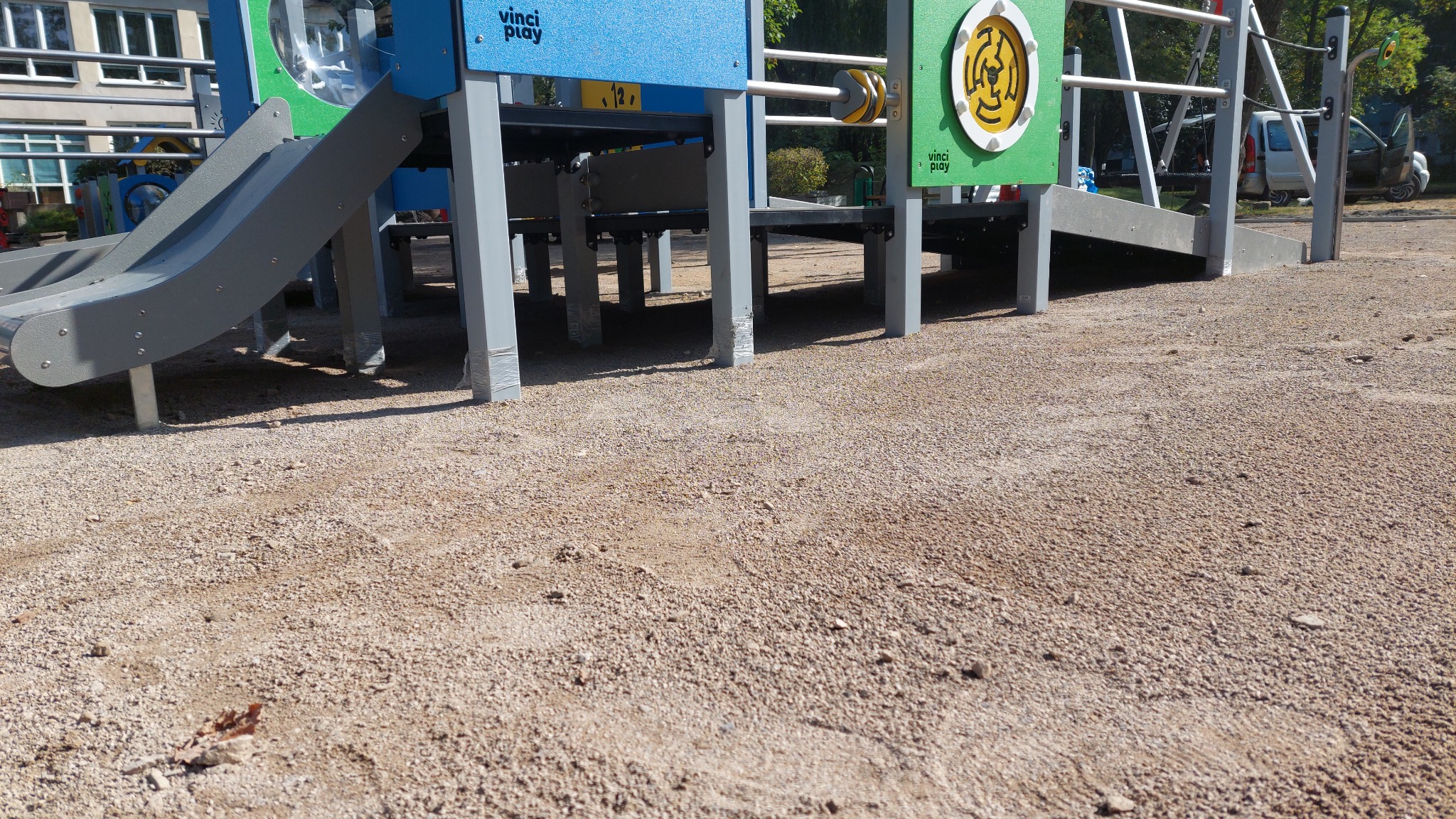Wieloelementowy plac zabaw z szarą zjeżdżalnią i elementami edukacyjnymi Vinci Play, otoczony żwirową nawierzchnią, widok z poziomu gruntu.