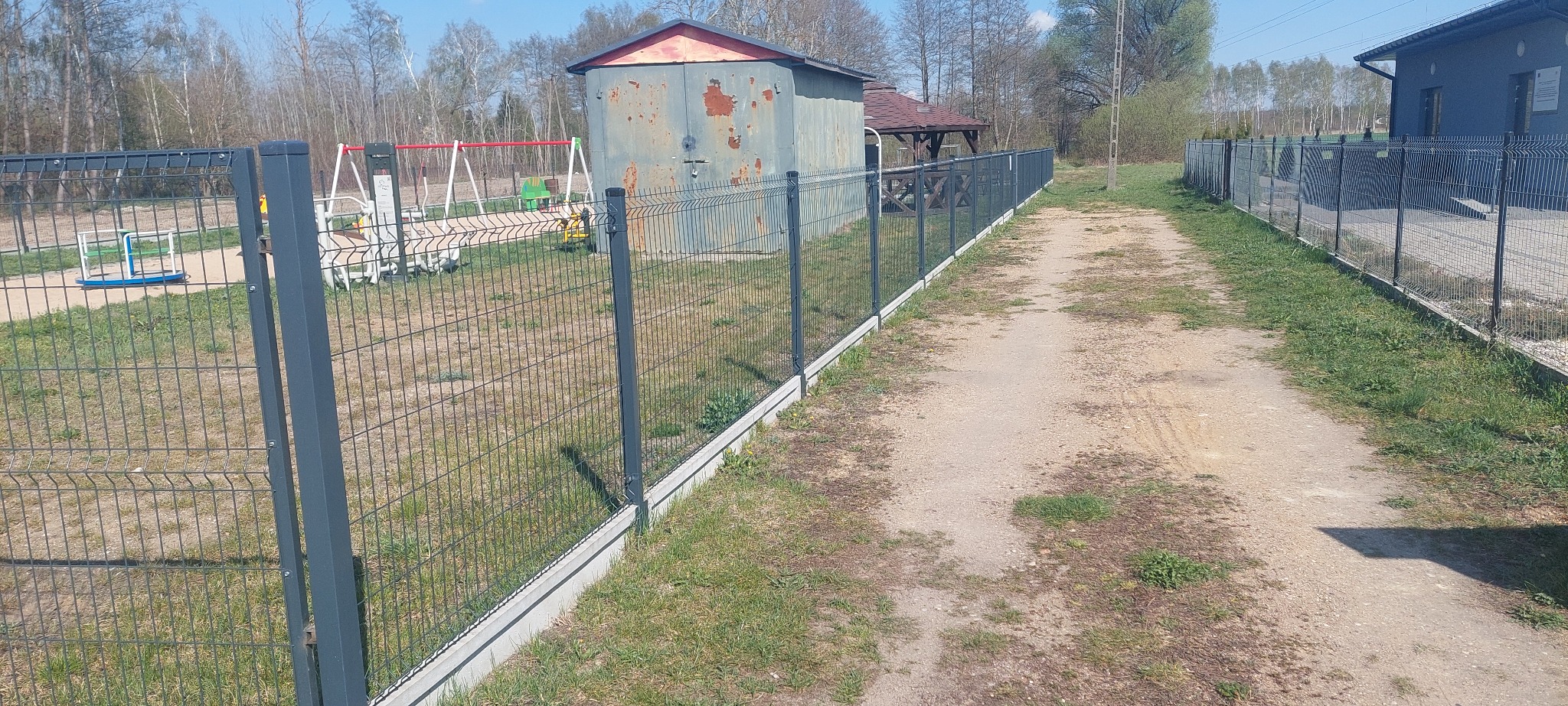 Panelowe ogrodzenie z podmurówką betonową, widok z boku na tle placu zabaw i zabudowań gospodarczych.