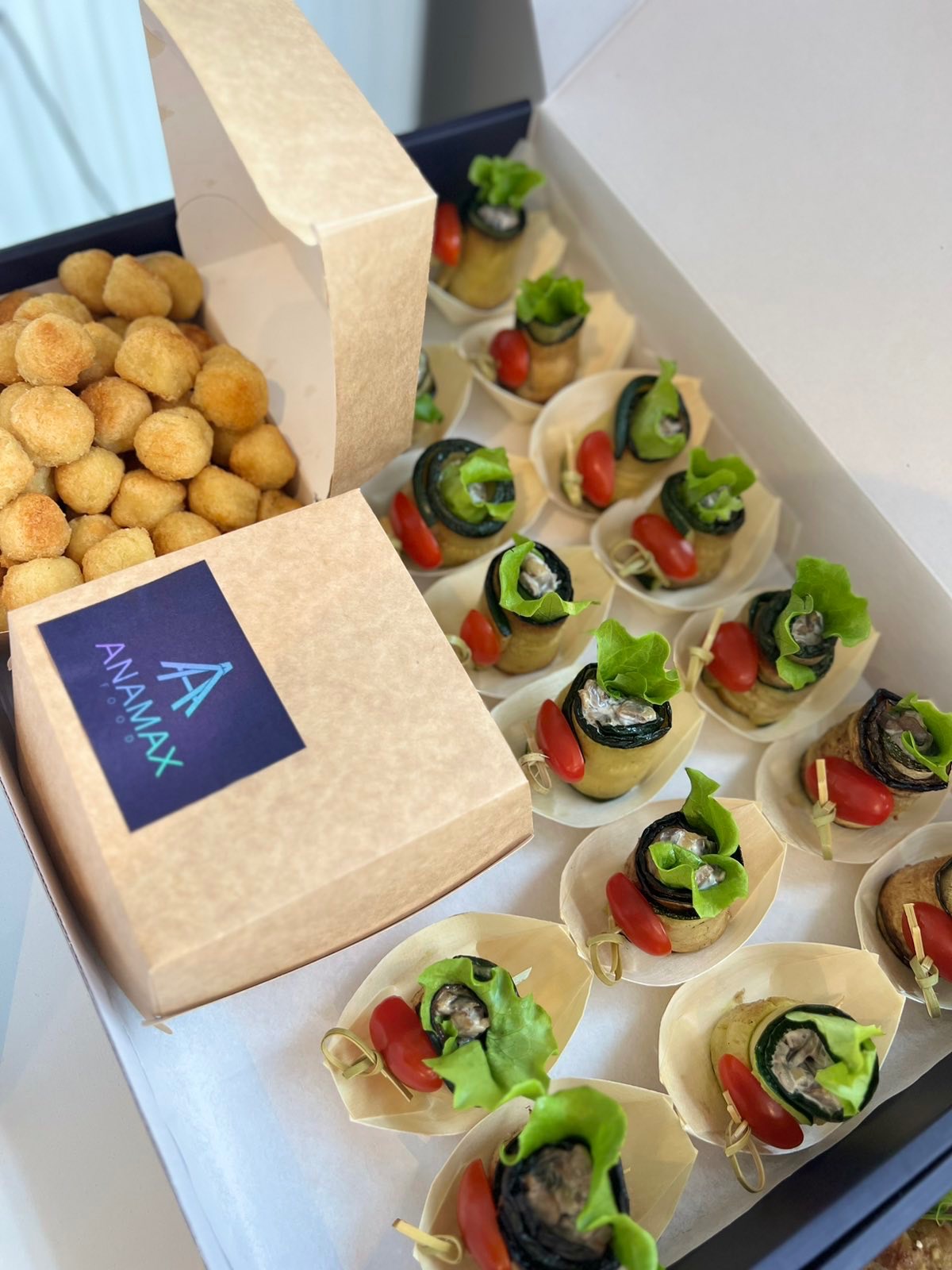 Elegancki catering: pudełko z roladkami z bakłażana, pomidorkami cherry i sałatą, obok pudełko z kulkami serowymi, całość z logo firmy Anamax.
