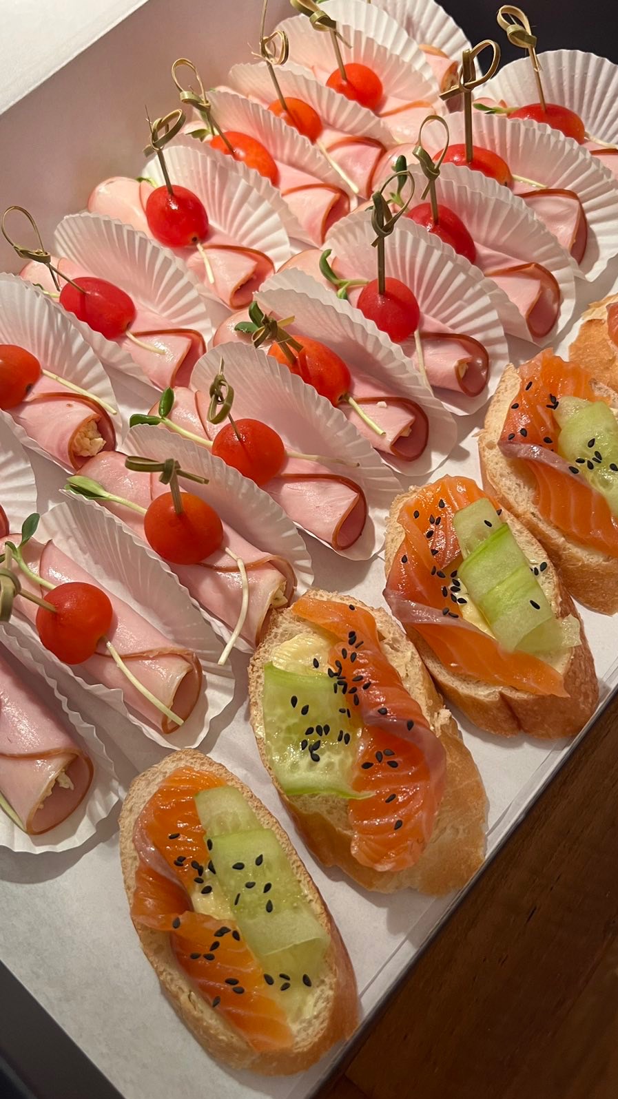 Elegancki catering: Roladki z szynki z pomidorkiem cherry i kanapki z łososiem, ogórkiem i czarnym sezamem, ułożone w białym pudełku.