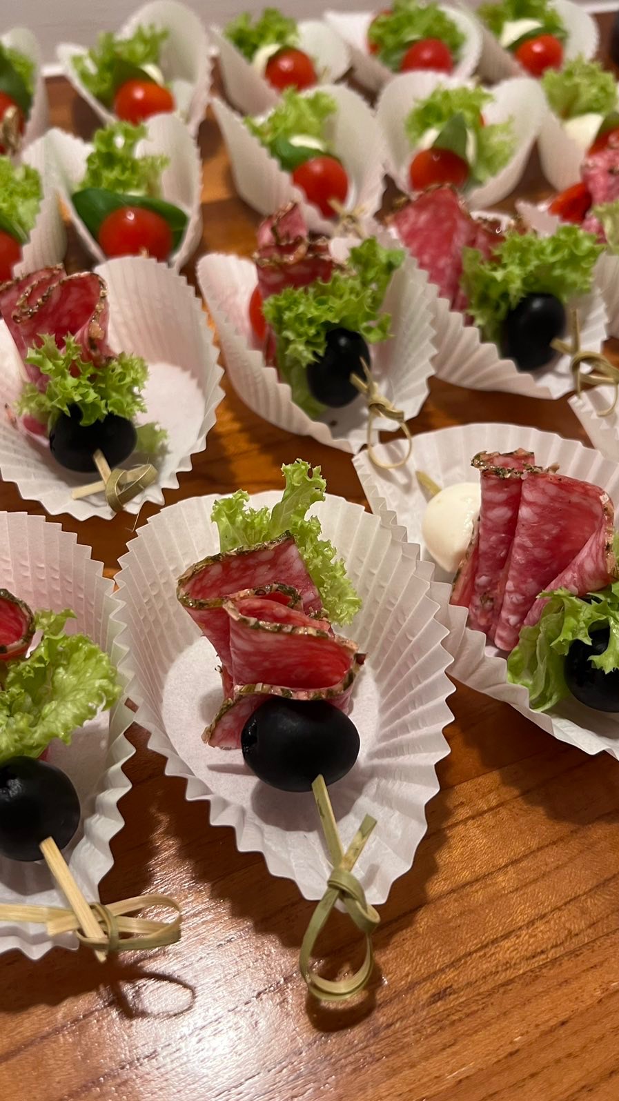 Apetyczne koreczki na drewnianym stole, każdy w papierowej foremce. Wędlina, oliwki, pomidorki i zielenina tworzą elegancką przekąskę.