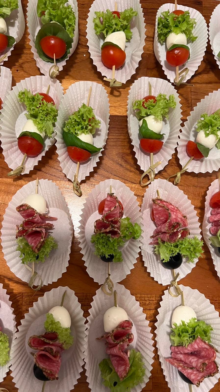 Dekoracyjne koreczki: mozzarella, pomidorki, salami i sałata na drewnianych szpadkach, ułożone w papierowych papilotkach na drewnianym stole. Elegancki catering.