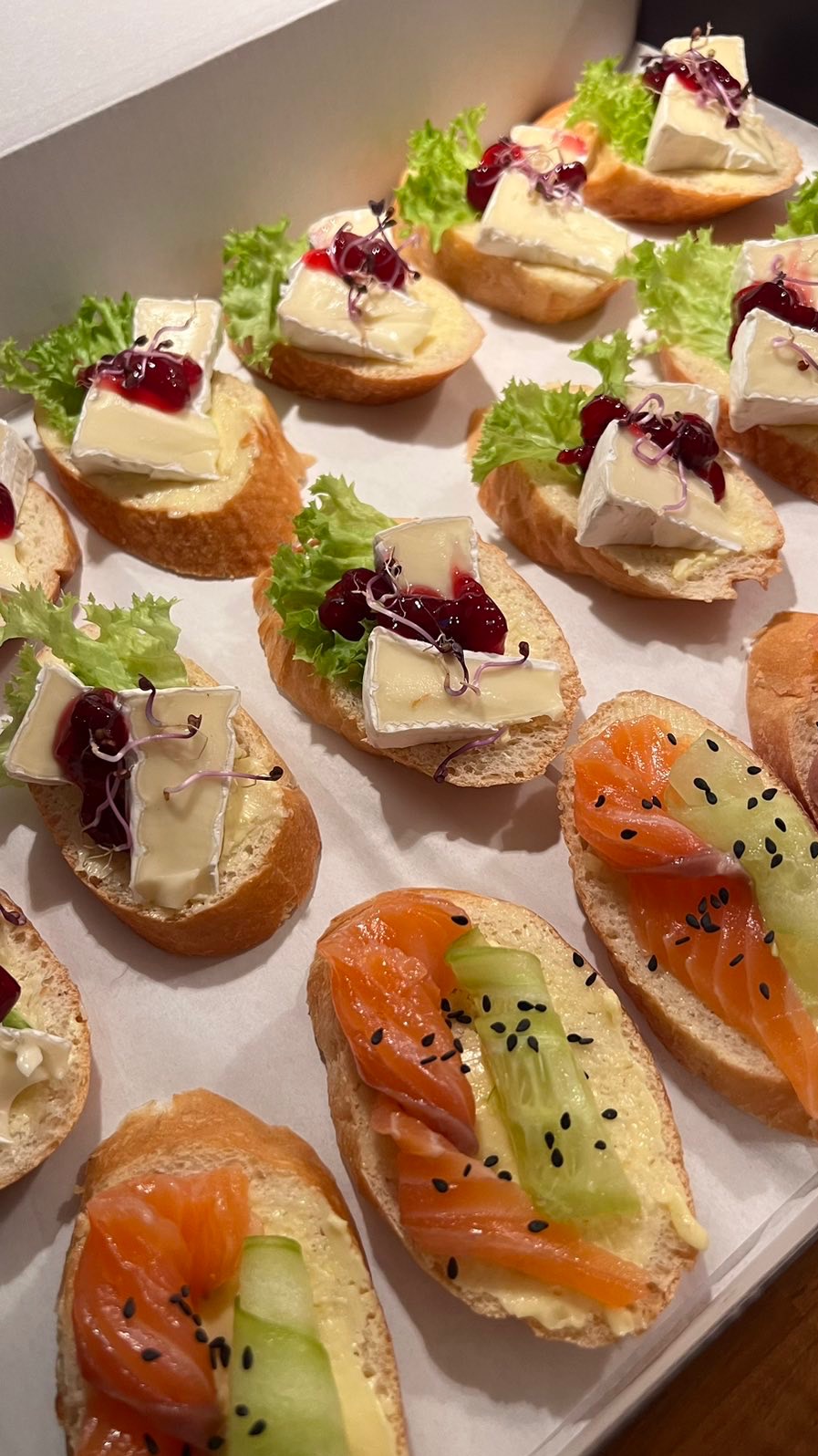 Apetyczne kanapeczki cateringowe: bagietki z brie, sałatą, żurawiną i łososiem z ogórkiem, posypane czarnym sezamem, ułożone na białym tle.