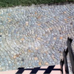 Wykończona powierzchnia z kostki granitowej ułożonej w koncentryczne okręgi, widoczne schody i trawnik w tle.