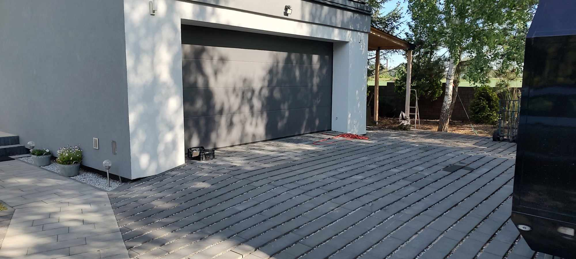 Ułożona, szara kostka brukowa przed nowoczesnym garażem z bramą segmentową, widoczny fragment elewacji domu, rabata z kwiatami w donicach i akcenty w postaci białego żwiru.