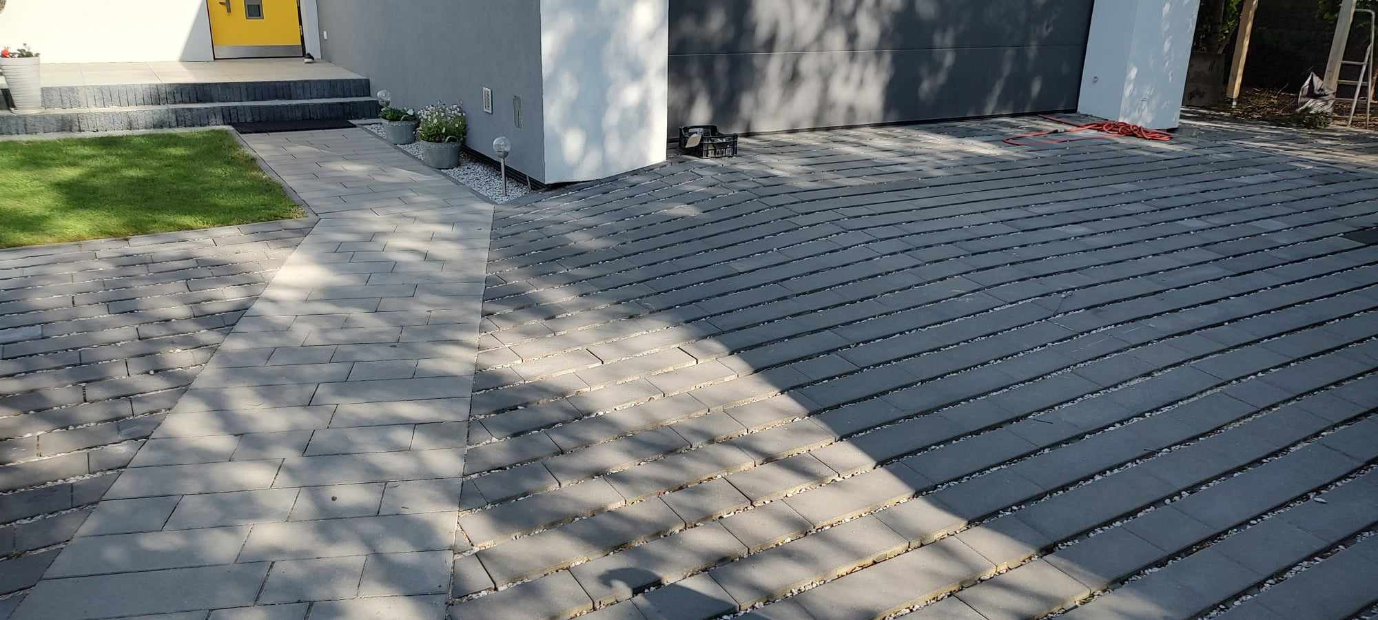 Nowoczesny podjazd z szarej kostki brukowej ułożonej w regularny wzór, z jasną kostką prowadzącą do drzwi wejściowych, uzupełniony białym żwirem w szczelinach i zielonym trawnikiem po lewej stronie.