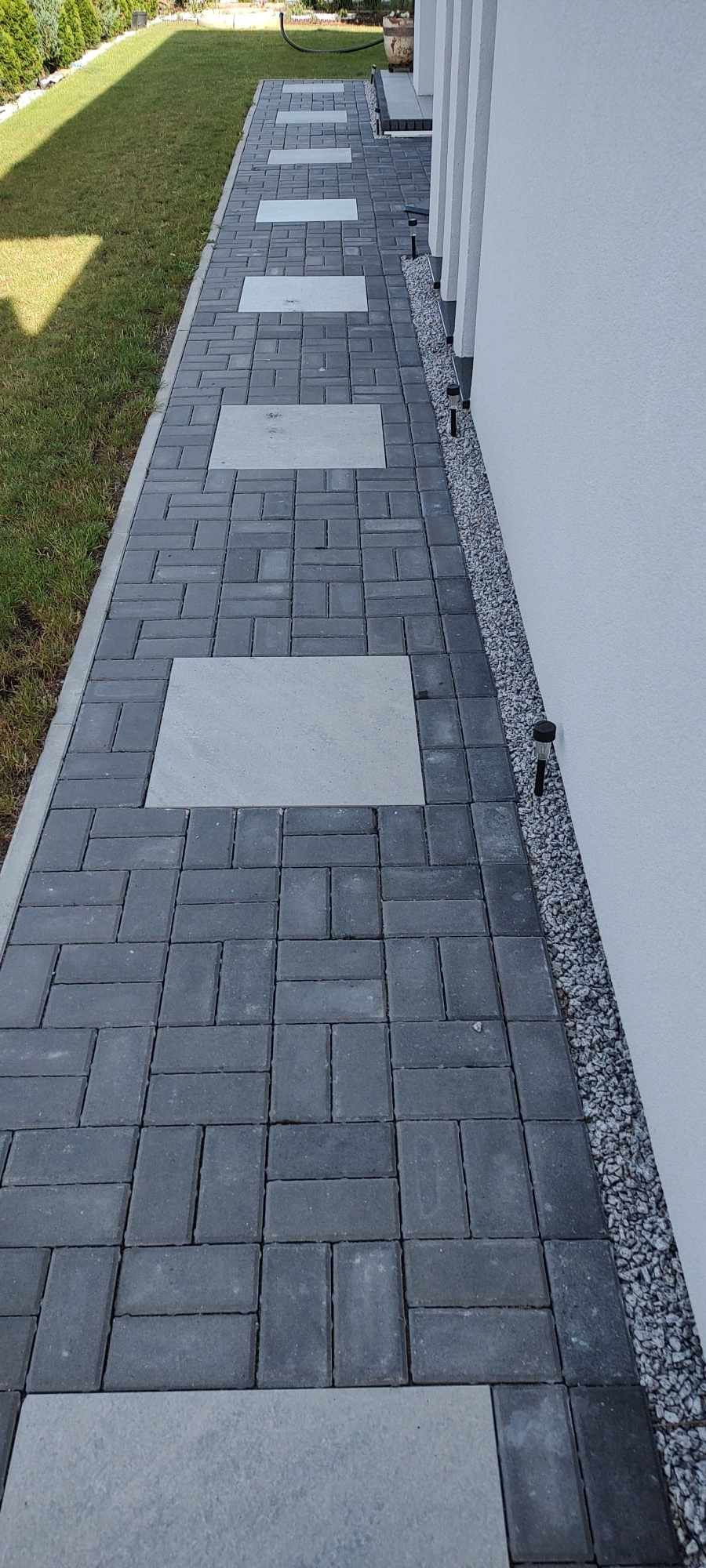 Ścieżka z ciemnoszarej kostki brukowej ułożonej w regularny wzór, z wstawkami z jasnoszarych płyt, prowadząca wzdłuż białej ściany budynku. Wzdłuż ściany pas drobnego żwiru, a po drugiej stronie...