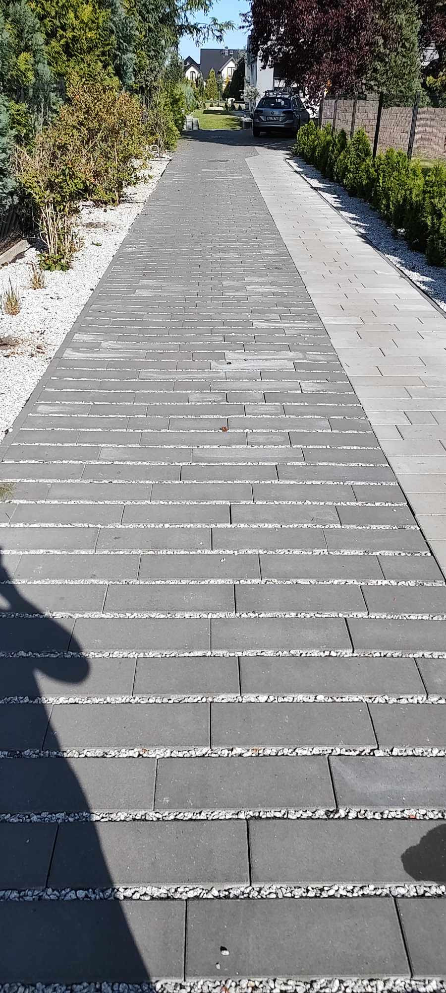 Długa, szara ścieżka z kostki brukowej, z białym żwirem między rzędami, prowadząca do domu z ogrodem w tle.