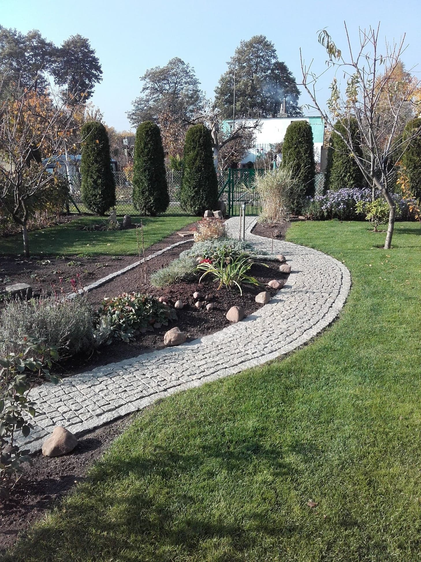 Kręta ścieżka z jasnej kostki granitowej w ogrodzie, obsadzona roślinami i otoczona trawnikiem, prowadząca do zielonej bramki.