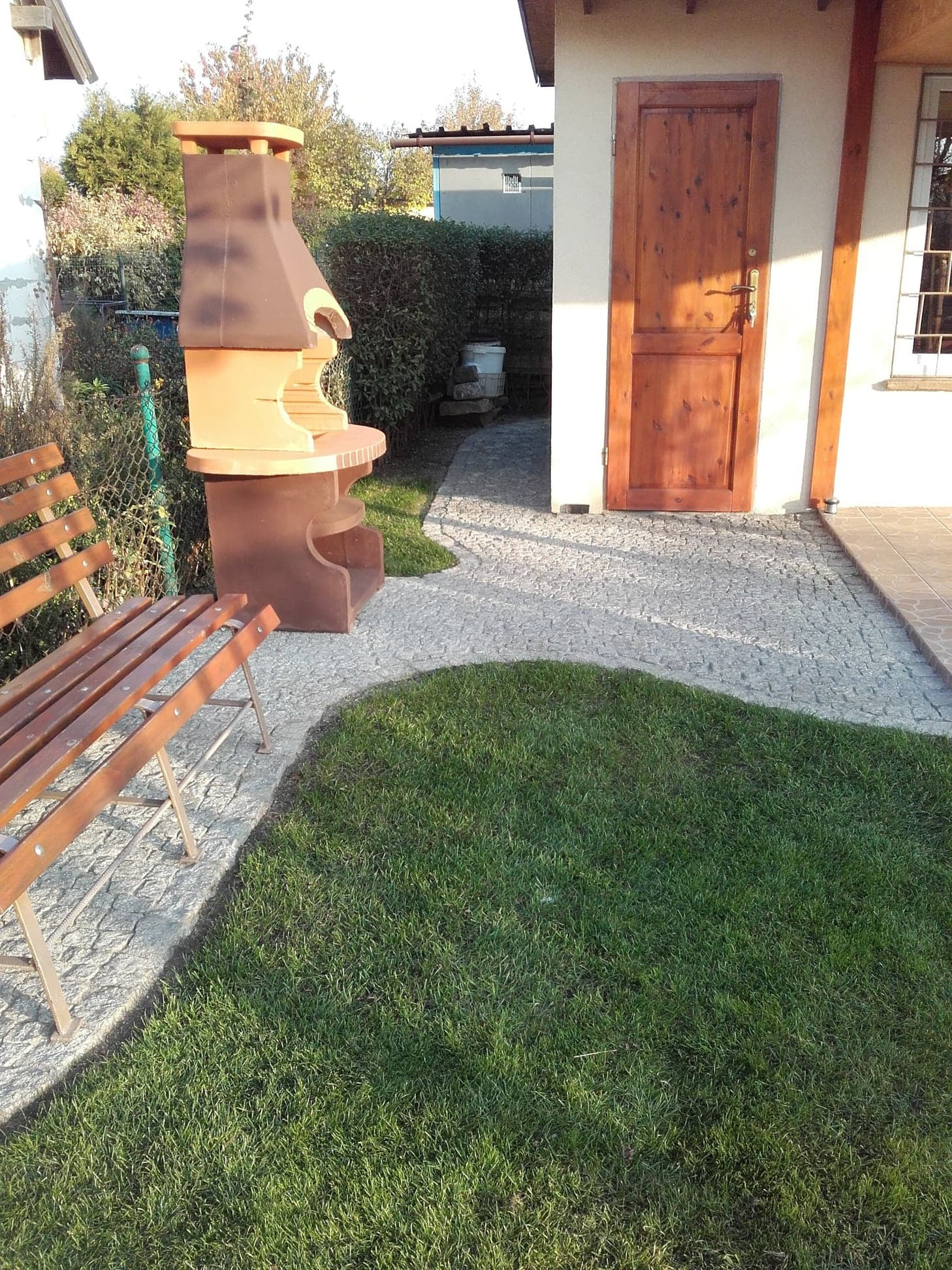 Ułożona ścieżka z jasnoszarej kostki granitowej prowadząca do drewnianych drzwi budynku gospodarczego, obok trawnik, drewniana ławka i murowany grill.