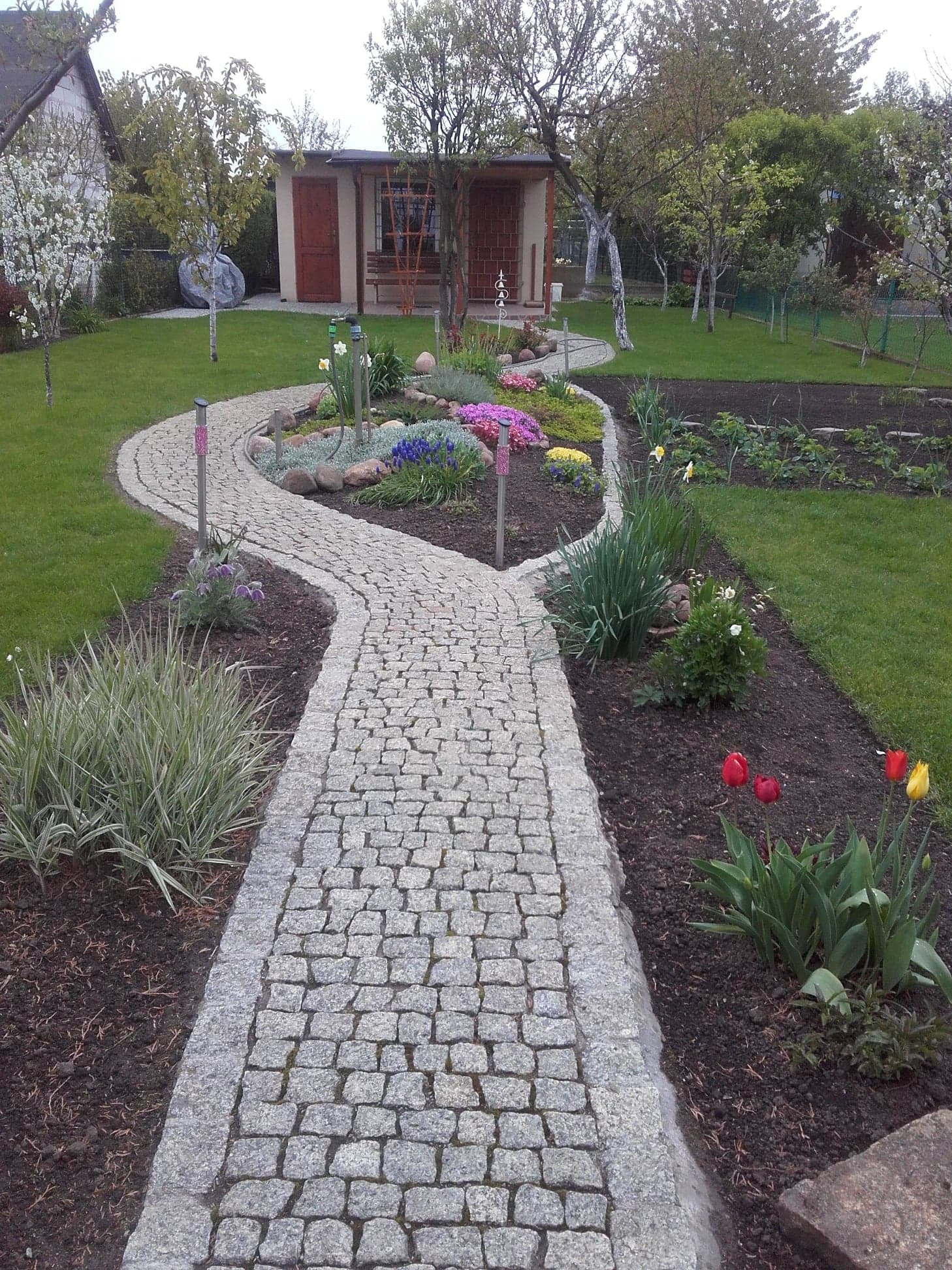 Kręty chodnik z jasnoszarej kostki granitowej prowadzący przez zadbany ogród z rabatami kwiatowymi, z widoczną altaną w tle.