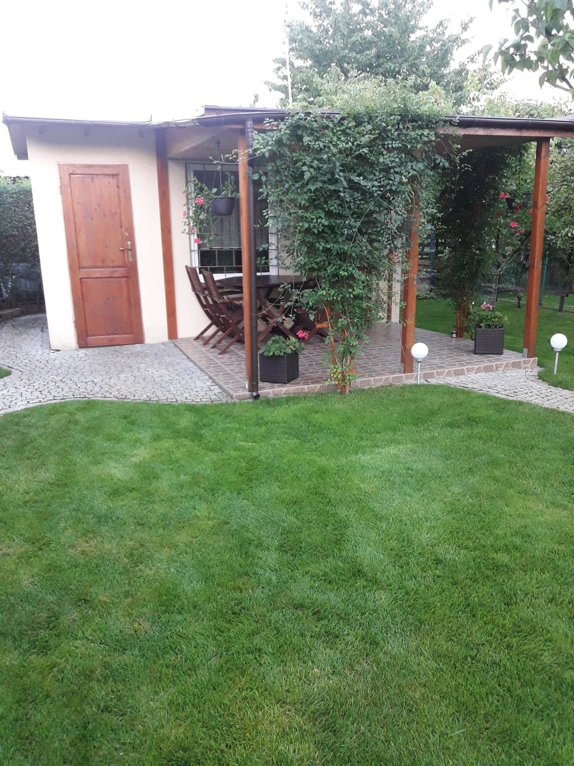 Ogród z altaną porośniętą pnączami, obok mały domek i ścieżka z kostki granitowej prowadząca przez trawnik.