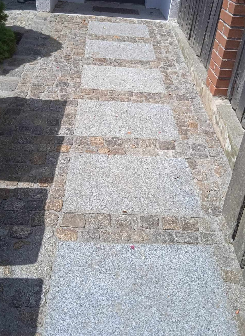 Ścieżka z płyt granitowych i kostki brukowej w odcieniach szarości i beżu, prowadząca do wejścia budynku, widoczny cień rzucany przez roślinność.