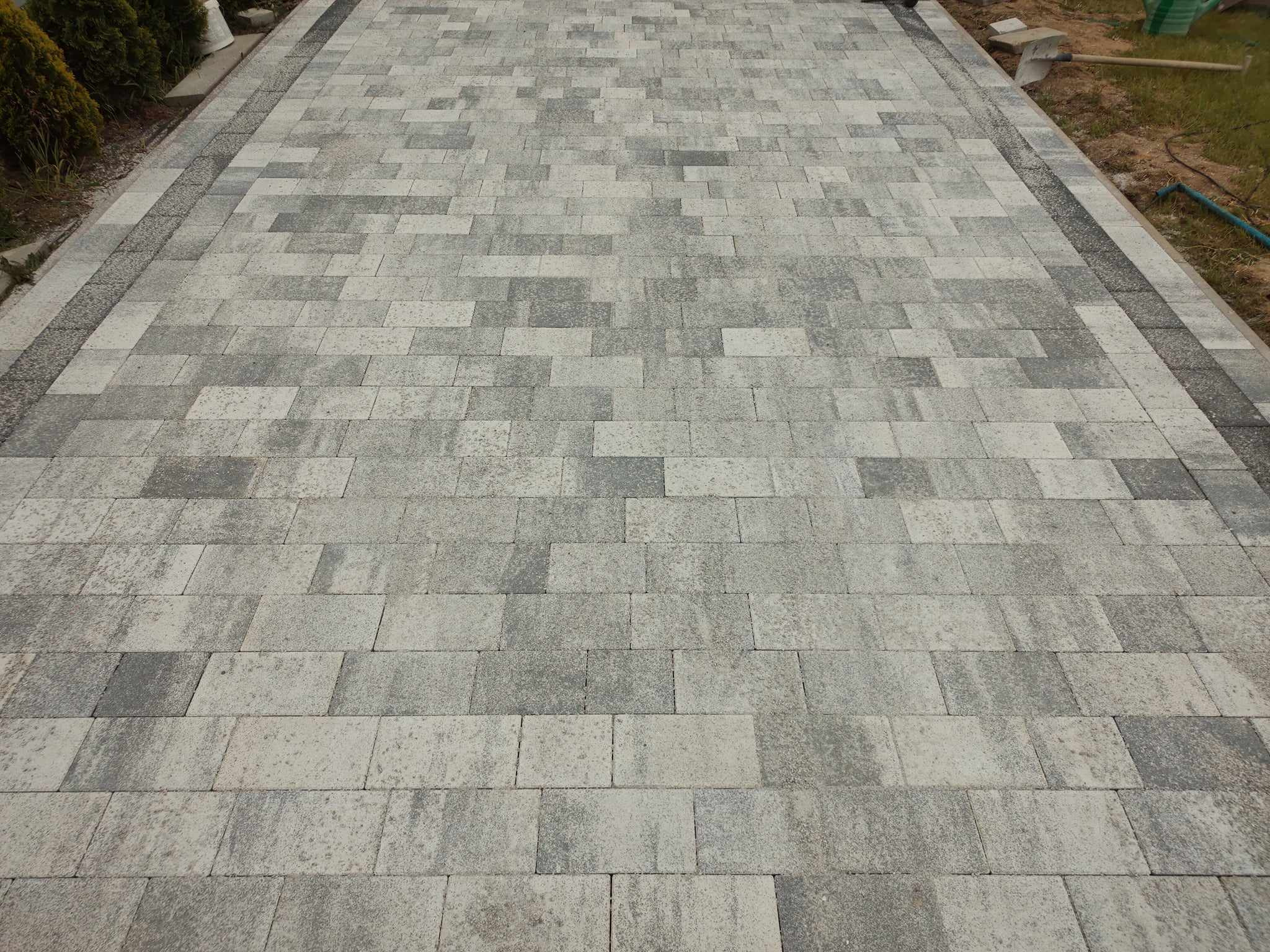 Ułożona ścieżka z kostki brukowej w odcieniach szarości, z ciemniejszym obramowaniem, widok z góry w perspektywie, z narzędziami w tle.