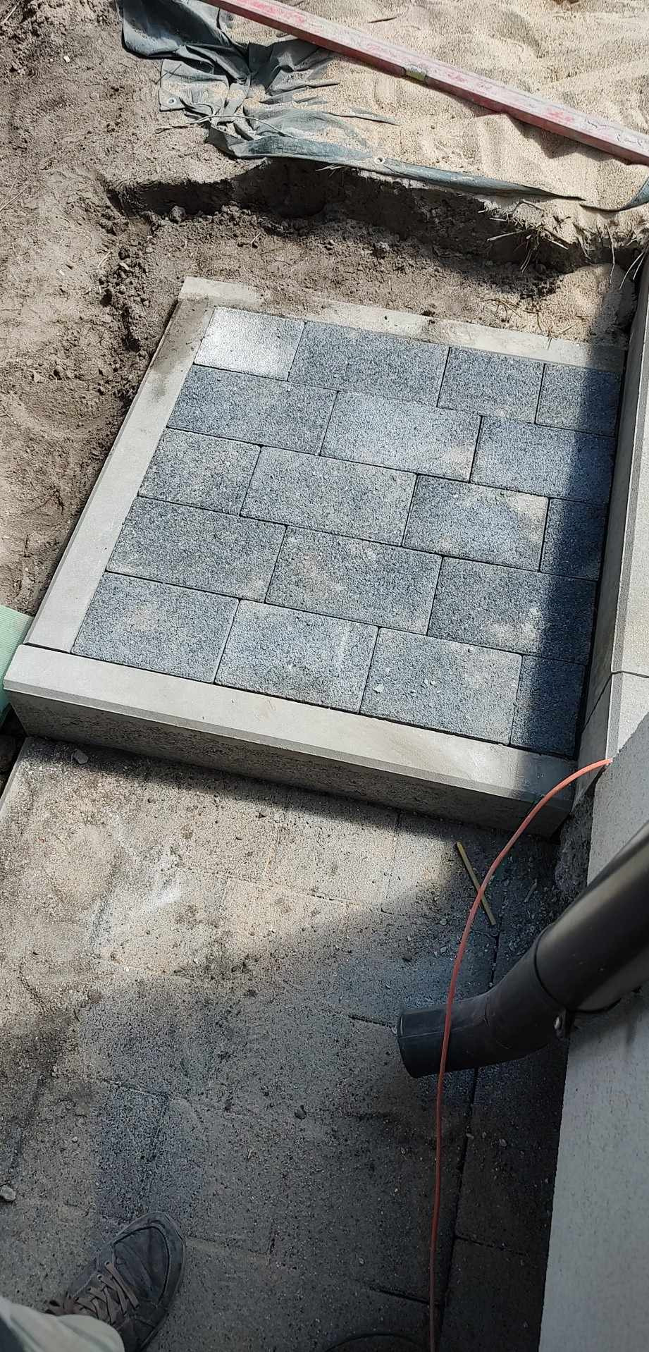 Fragment nowo ułożonej, szarej kostki brukowej z obramowaniem, widoczny wykop pod dalszą część nawierzchni.