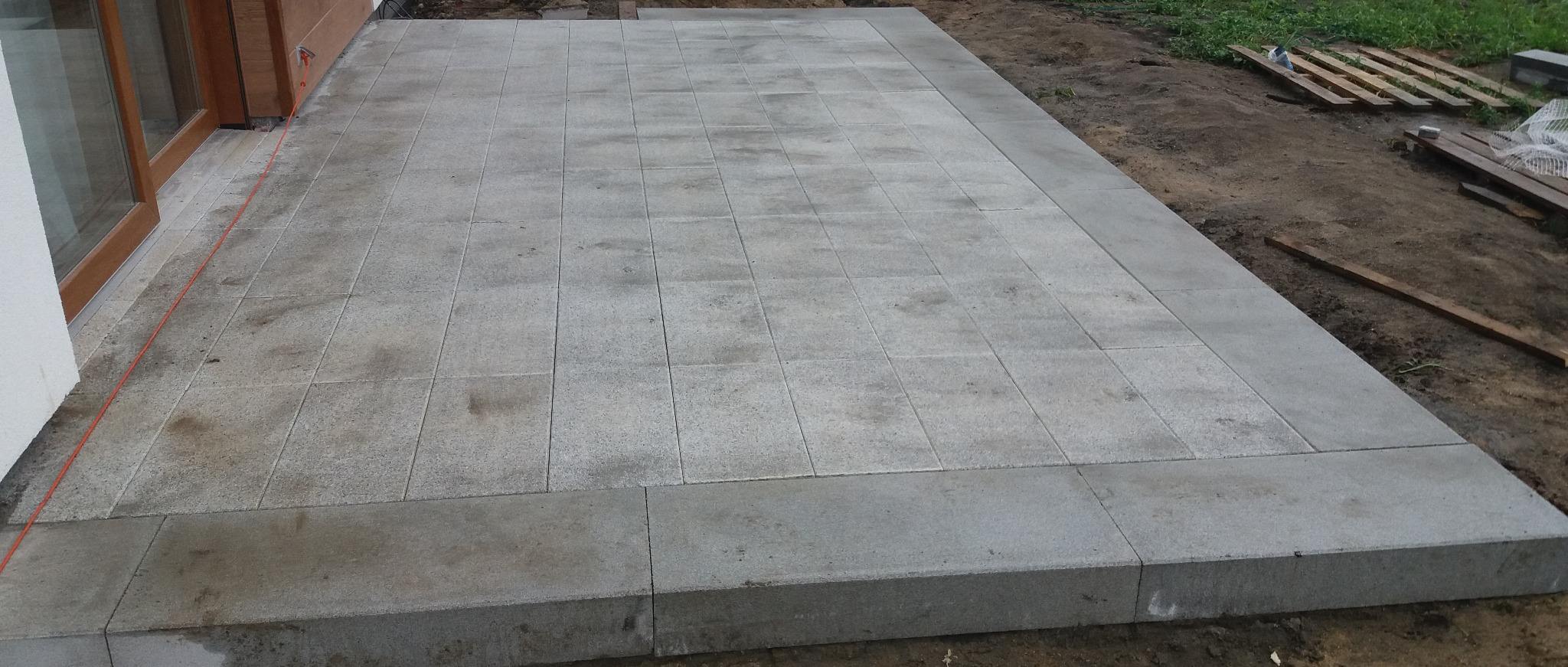 Szara, ułożona kostka na tarasie przy budynku, z widoczną czerwoną linką wyznaczającą linię, na ziemi obok leżą drewniane palety.